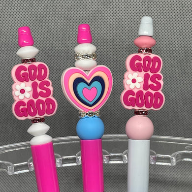 Three decorative pens with 'God is Good' text and heart design on a gray background